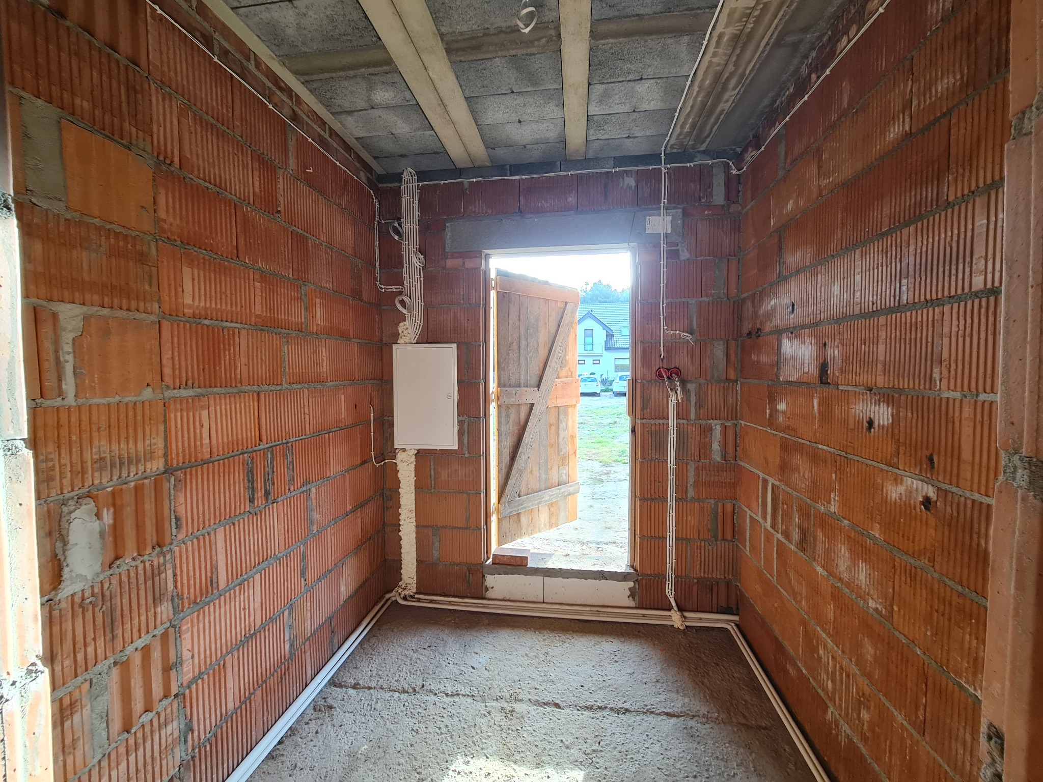 Wnętrze w trakcie budowy z widoczną instalacją elektryczną na ścianach z czerwonej cegły. Skrzynka elektryczna przy otwartych drzwiach. Widoczne przewody i rury.