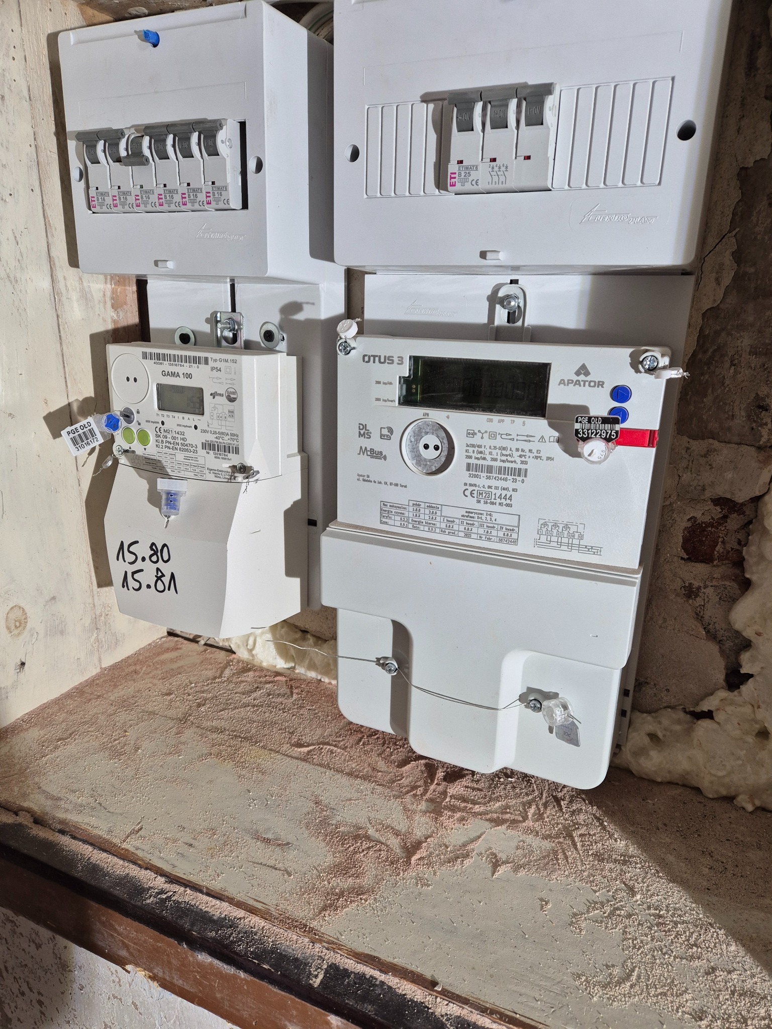 Dwa liczniki elektryczne (GAMA 100 i OTUS 3) zamontowane obok skrzynki z bezpiecznikami wewnątrz budynku. Widoczne oznaczenia i numery seryjne urządzeń.