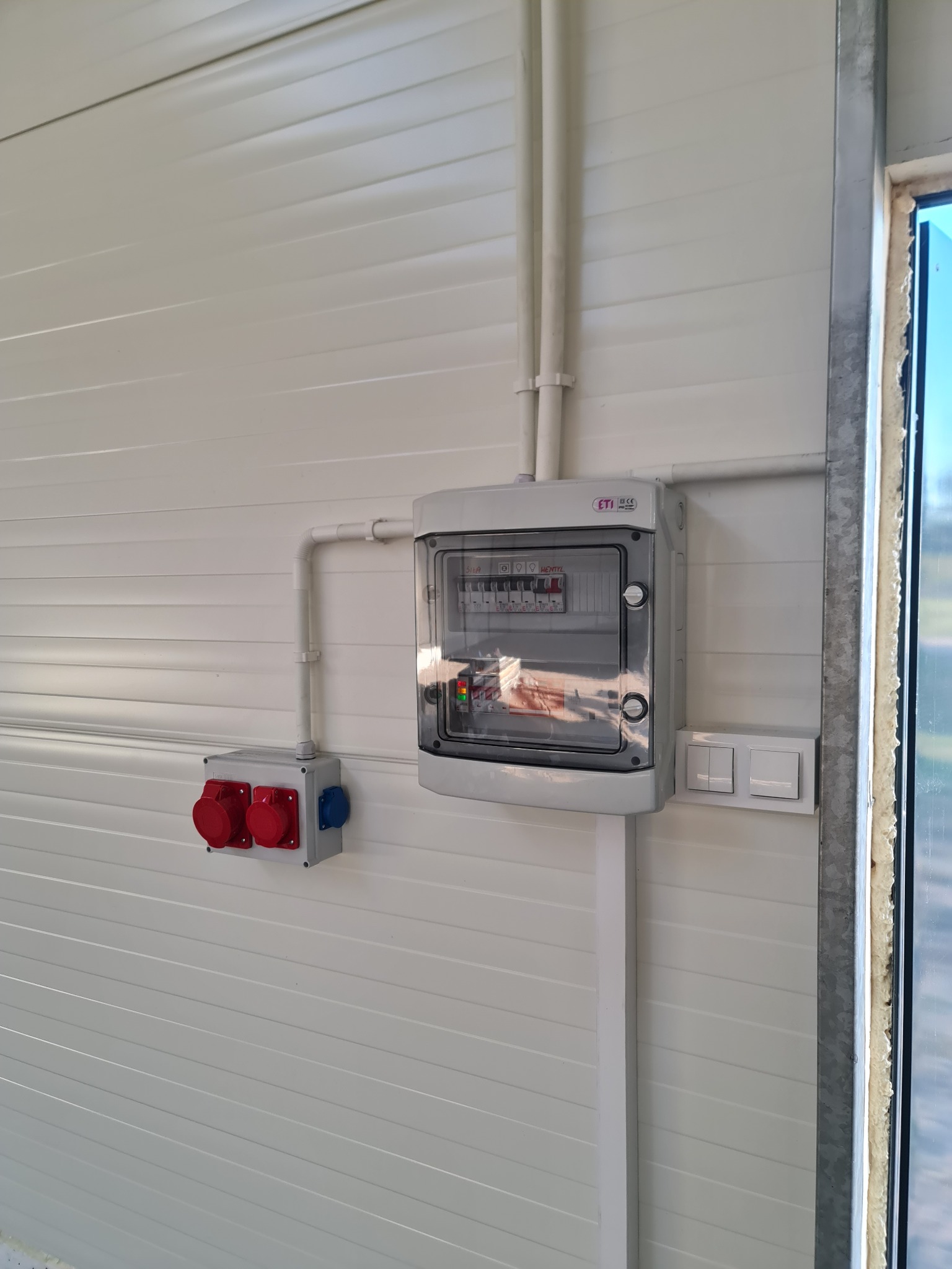 Szara skrzynka rozdzielcza z bezpiecznikami, gniazda siłowe (czerwone i niebieskie) oraz włączniki światła na białej ścianie z paneli. Widoczne rury instalacyjne.