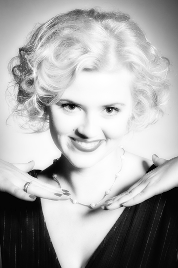 Portret kobiety z blond, pofalowaną fryzurą, w czarnej sukience z dekoltem w serek, prezentującej delikatny naszyjnik z pereł, z dłońmi ułożonymi wokół szyi, w czarno-białej tonacji.