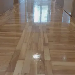 Błyszcząca, świeżo polakierowana podłoga parkietowa z jasnego drewna, ułożona w pasy, odbijająca światło okna w długim korytarzu z beżowymi ścianami.