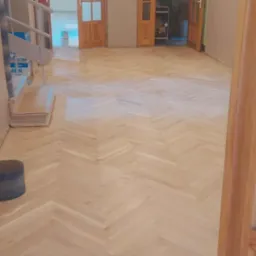 Jasny parkiet ułożony w jodełkę, widoczny z perspektywy otwartych, drewnianych drzwi z szybami. W tle schody i kolejne pomieszczenia.