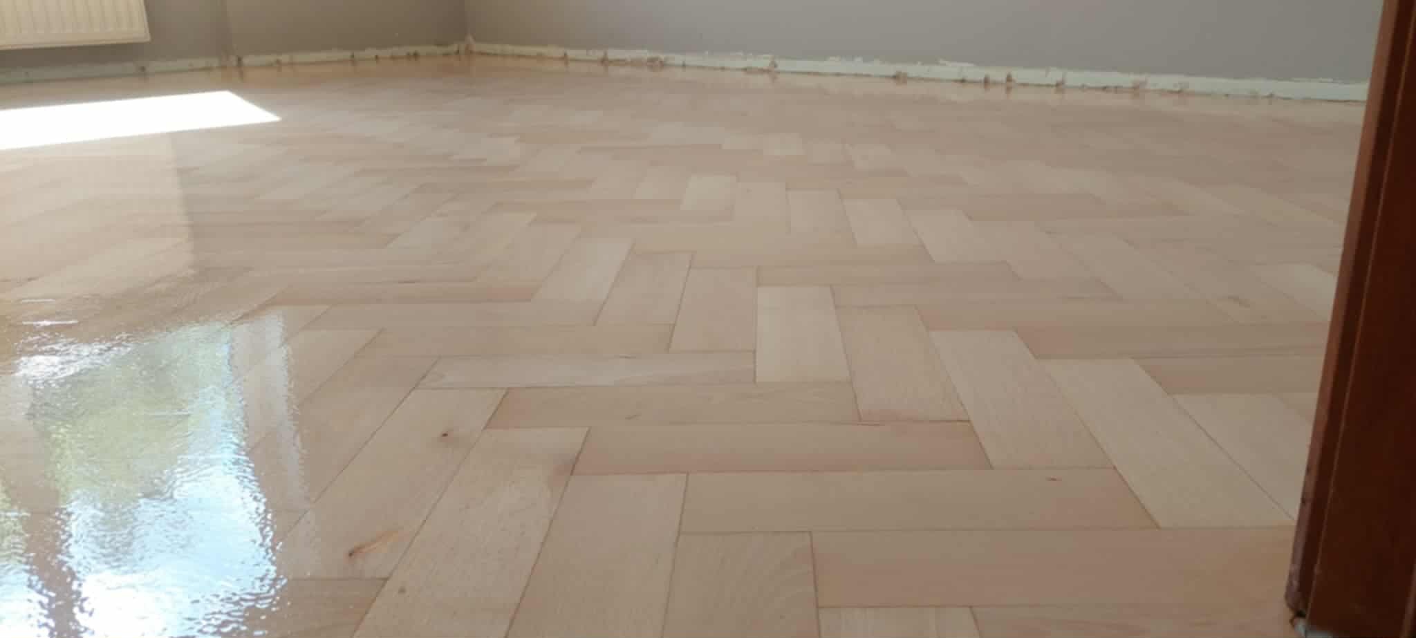 Jasny parkiet ułożony w jodełkę, świeżo polakierowany, z widocznym odbiciem światła. Listwy przypodłogowe w kolorze ściany. Fragment drzwi po prawej stronie kadru.