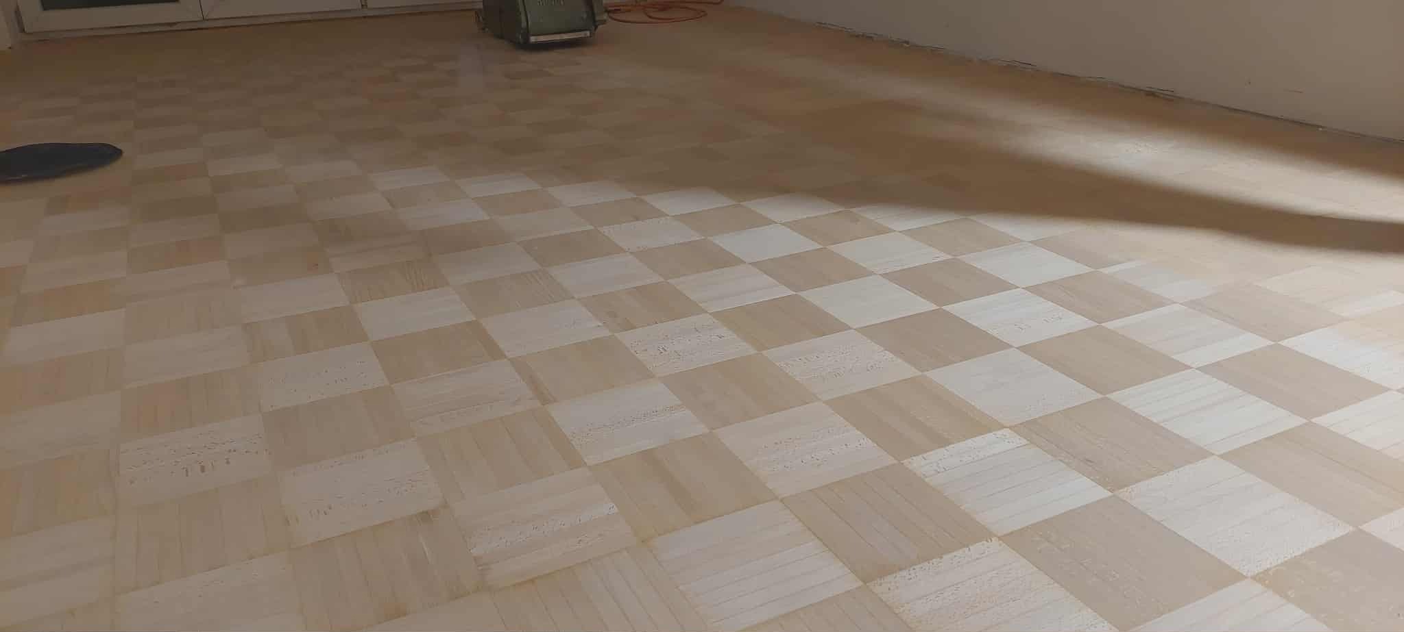 Szachownicowy parkiet po renowacji, z widocznym sprzętem do cyklinowania w tle. Jasne drewno, świeżo po obróbce, z delikatnym połyskiem. Widok z góry, równomierne oświetlenie.