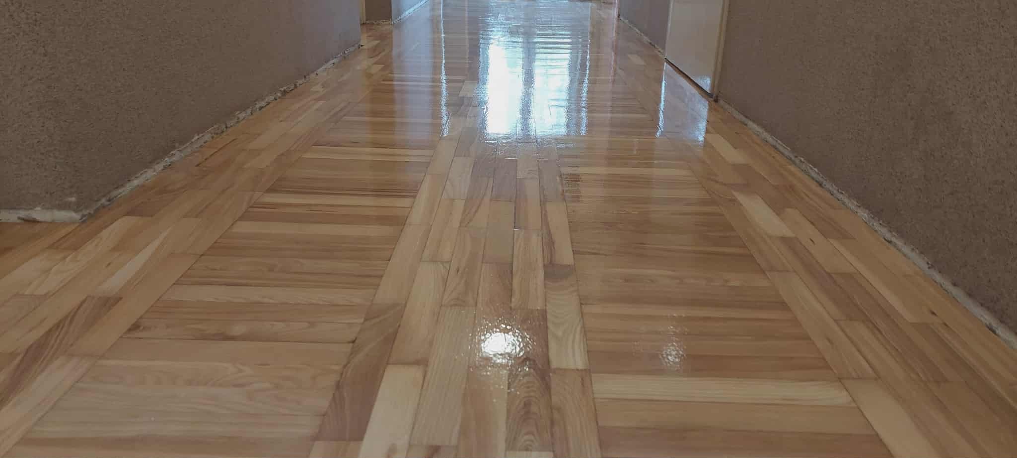 Błyszcząca, świeżo polakierowana podłoga parkietowa z jasnego drewna, ułożona w pasy, odbijająca światło okna w długim korytarzu z beżowymi ścianami.