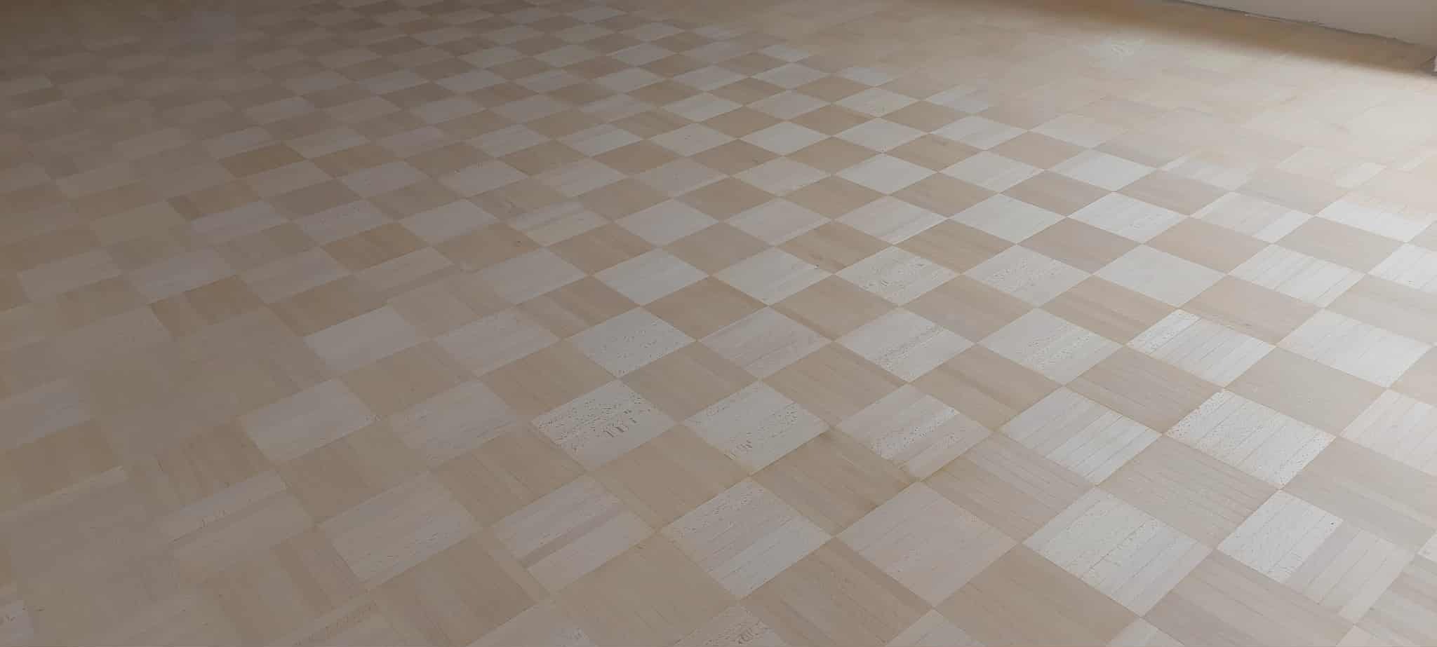 Szachownica z jasnego parkietu, świeżo po renowacji. Widoczne delikatne różnice w odcieniach drewna, podkreślające naturalny charakter podłogi. Minimalistyczne wnętrze.