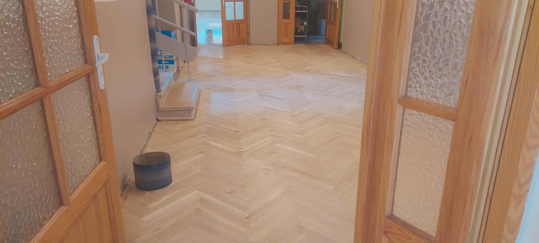 Jasny parkiet ułożony w jodełkę, widoczny z perspektywy otwartych, drewnianych drzwi z szybami. W tle schody i kolejne pomieszczenia.