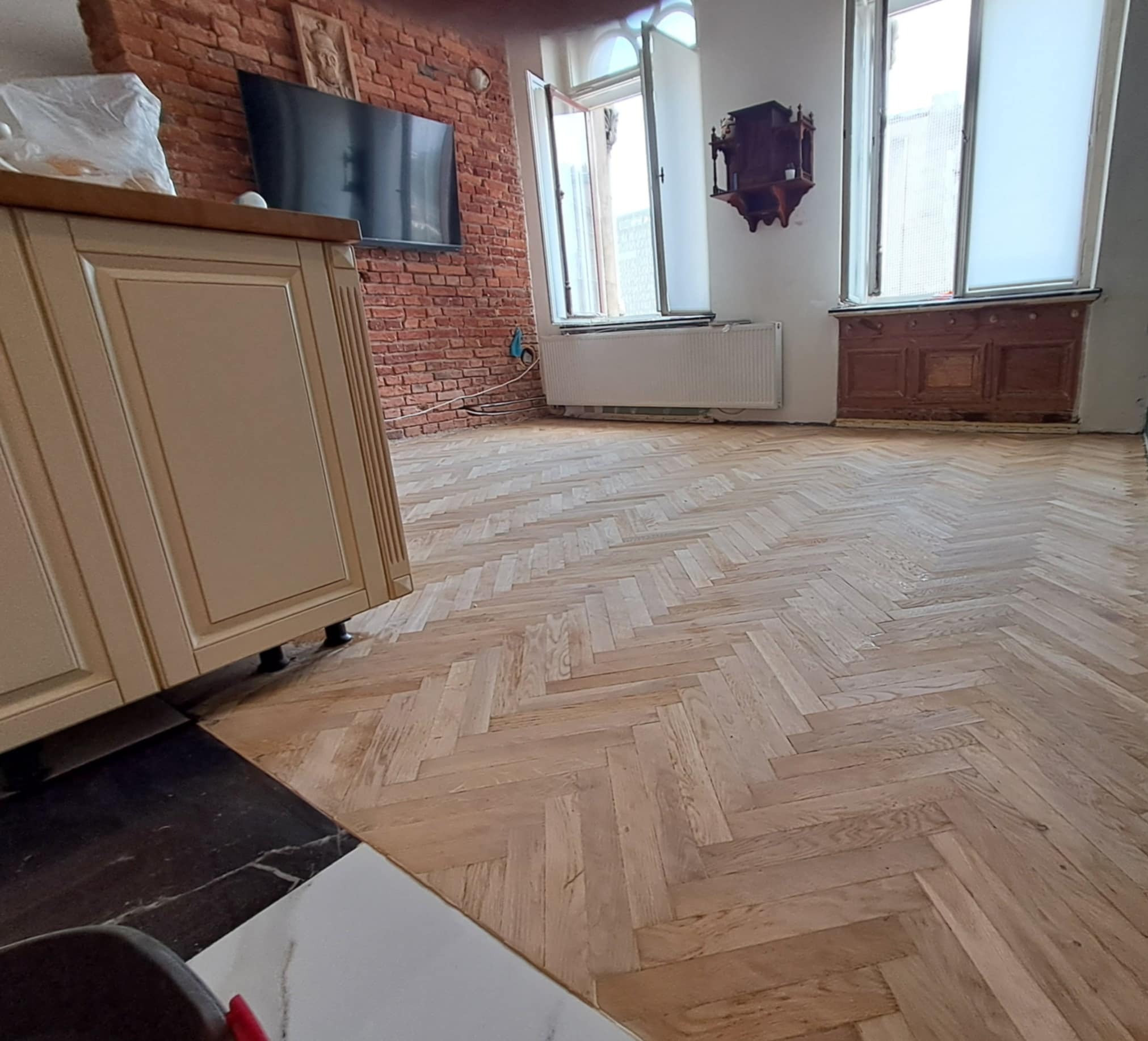 Jasny parkiet ułożony w jodełkę francuską w przestronnym wnętrzu z ceglaną ścianą i oknami. Widoczny fragment zabudowy kuchennej.