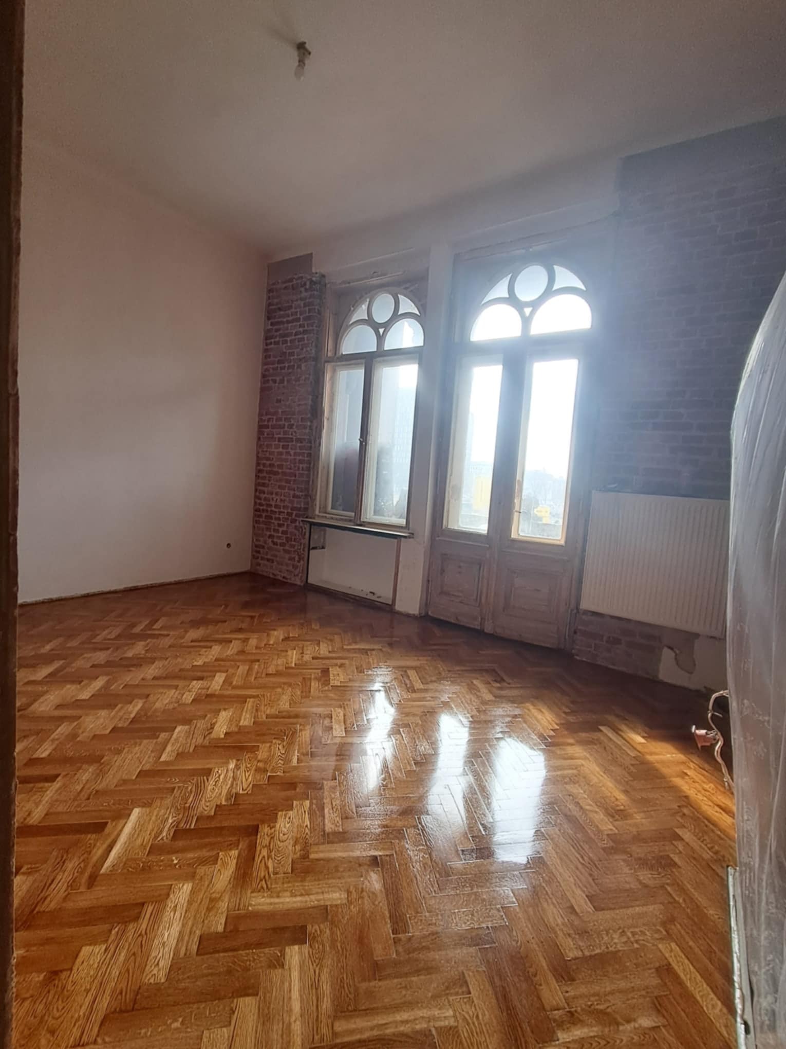 Błyszczący, odnowiony parkiet ułożony w jodełkę w przestronnym pokoju z ceglanymi wnękami okiennymi i starymi, drewnianymi oknami. Widoczne ślady renowacji.