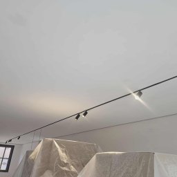 Adrian Rodak - Minimalistyczne oświetlenie szynowe z reflektorami punktowymi na białym suficie. Widoczne zabezpieczone meble folią malarską i okno w tle. Nowoczesny design.