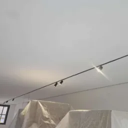 Minimalistyczne oświetlenie szynowe z reflektorami punktowymi na białym suficie. Widoczne zabezpieczone meble folią malarską i okno w tle. Nowoczesny design.