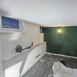 Adrian Rodak - Pokój w trakcie malowania: ściana pomalowana na zielono, białe ściany, okno, narzędzia na półce, podłoga zabezpieczona folią. Widoczne gniazdka elektryczne.