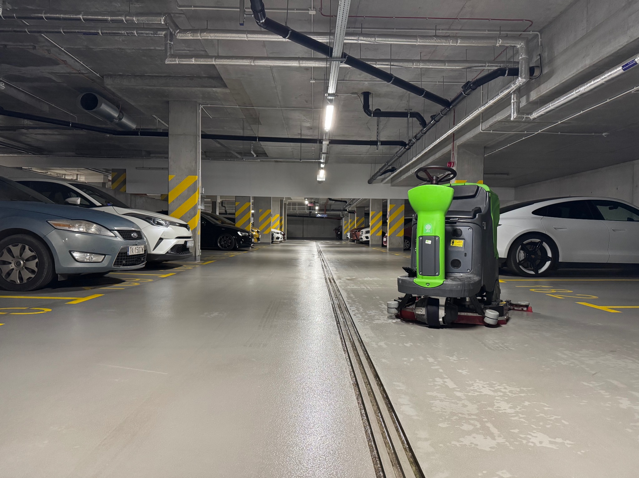 Profesjonalna maszyna czyszcząca w akcji na parkingu podziemnym, z widocznymi śladami wilgoci po myciu posadzki. W tle zaparkowane samochody.