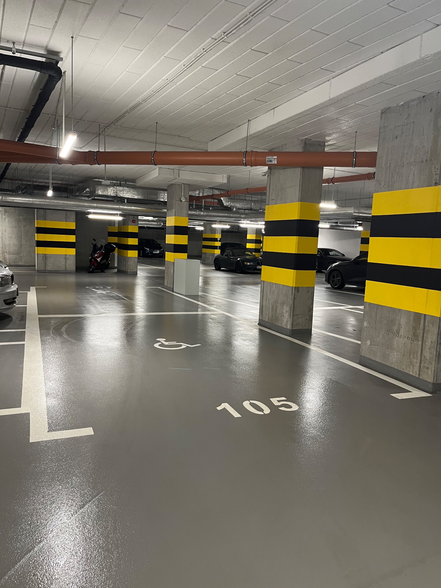 Błyszcząca, szara posadzka parkingu podziemnego z numerem 105 i symbolem osoby niepełnosprawnej. Żółto-czarne pasy na filarach, w tle zaparkowane samochody i motocykl.