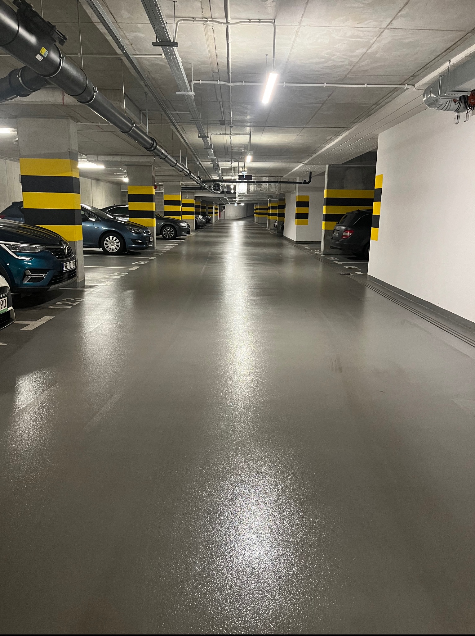 Błyszcząca, szara posadzka w garażu podziemnym z rzędami zaparkowanych aut i słupami w czarno-żółte pasy. Widok w głąb garażu, oświetlenie sufitowe.