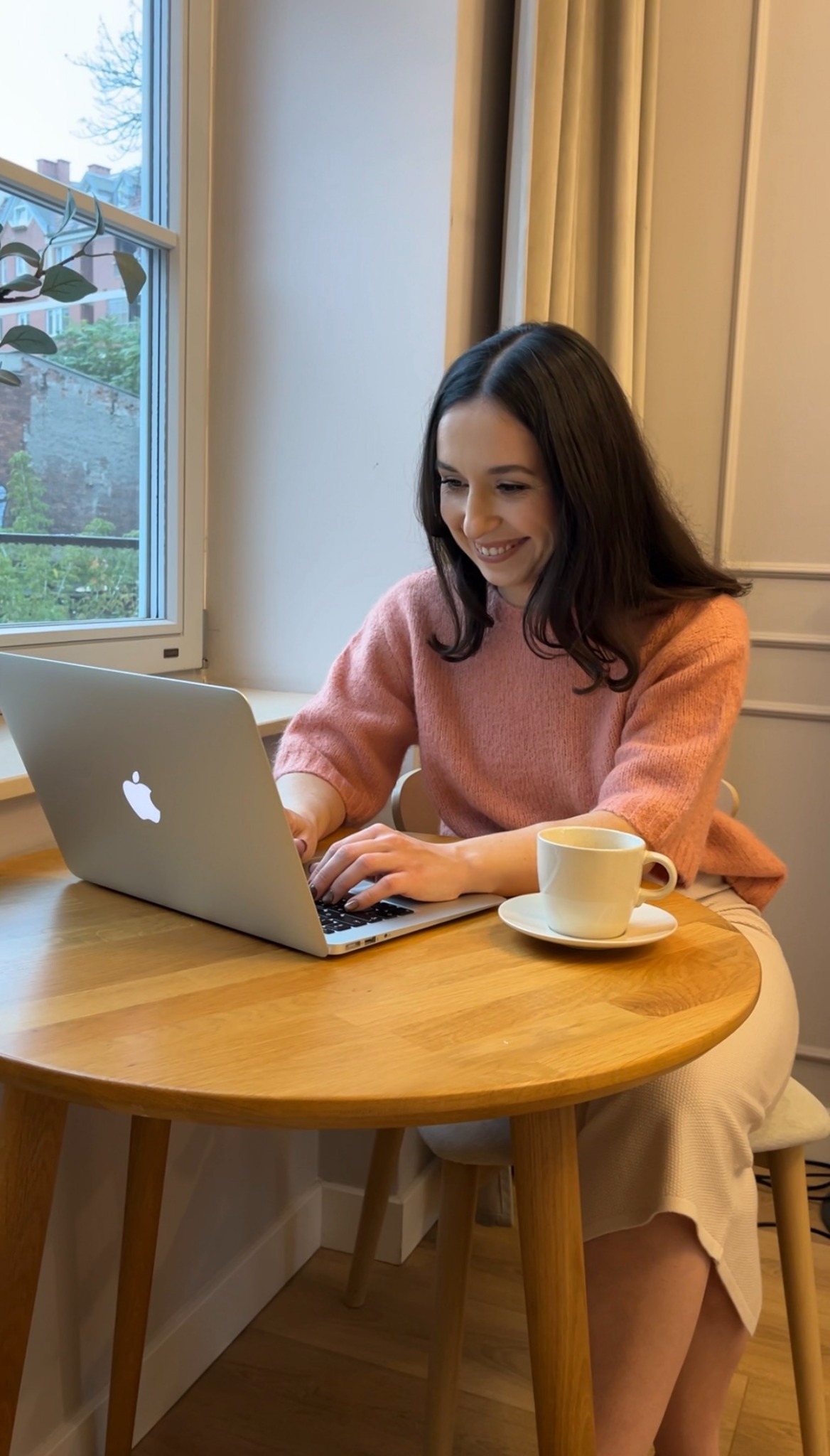 Uśmiechnięta kobieta w różowym swetrze pracuje na laptopie przy okrągłym drewnianym stole. Obok stoi filiżanka kawy. W tle okno z widokiem na budynek.
