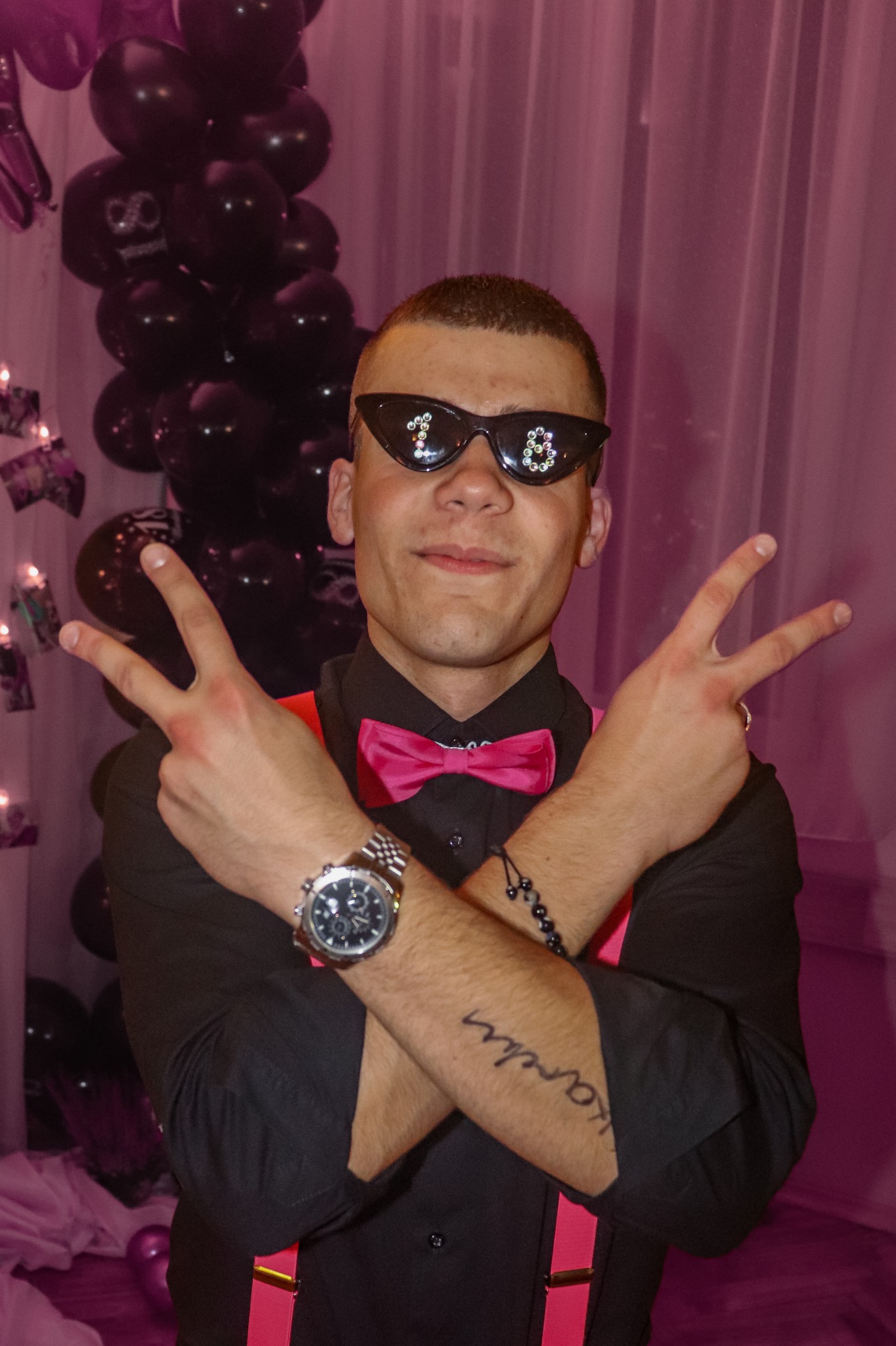 Młody mężczyzna w okularach z cyfrą 18, różowej muszce i szelkach, pozuje na tle balonów i zasłon. Uśmiech, gest zwycięstwa.