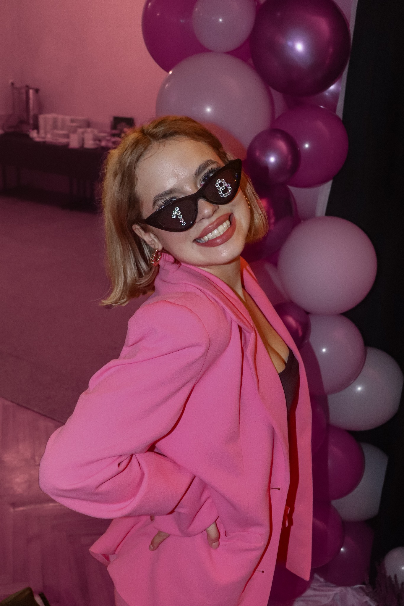 Uśmiechnięta blondynka w różowym żakiecie i okularach z cyrkoniami na tle różowych balonów. Zdjęcie z imprezy okolicznościowej, oświetlenie sceniczne.
