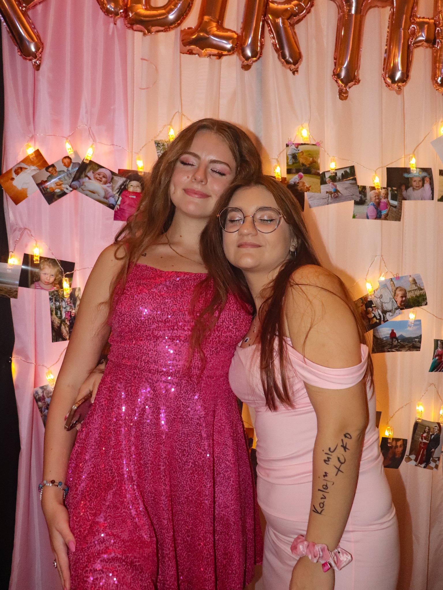 Dwie uśmiechnięte kobiety w różowych sukienkach pozują na tle różowej zasłony z balonami 'BIRTHDAY' i zdjęciami, oświetlonymi lampkami.