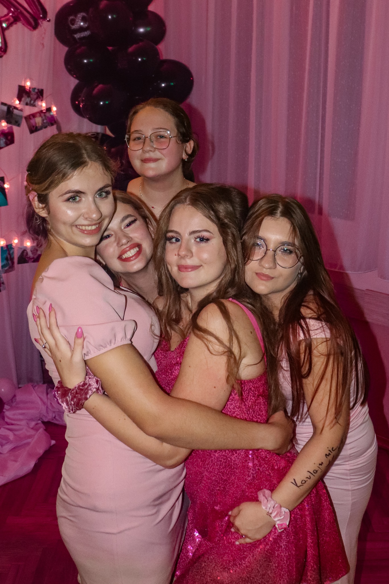 Grupowe portretowe ujęcie pięciu uśmiechniętych nastolatek w różowych sukienkach, obejmujących się na tle różowych dekoracji i balonów. Jedna z dziewcząt ma napis na ramieniu.