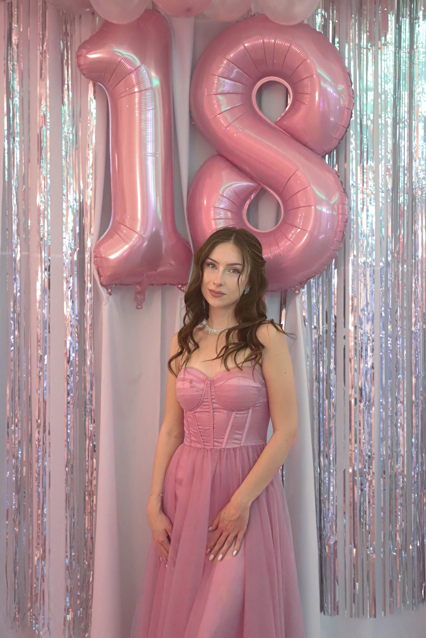 Portret młodej kobiety w różowej sukni na tle srebrnych frędzli i balonów w kształcie cyfr 1 i 8, świętującej osiemnaste urodziny. Uśmiech, delikatny makijaż i biżuteria.
