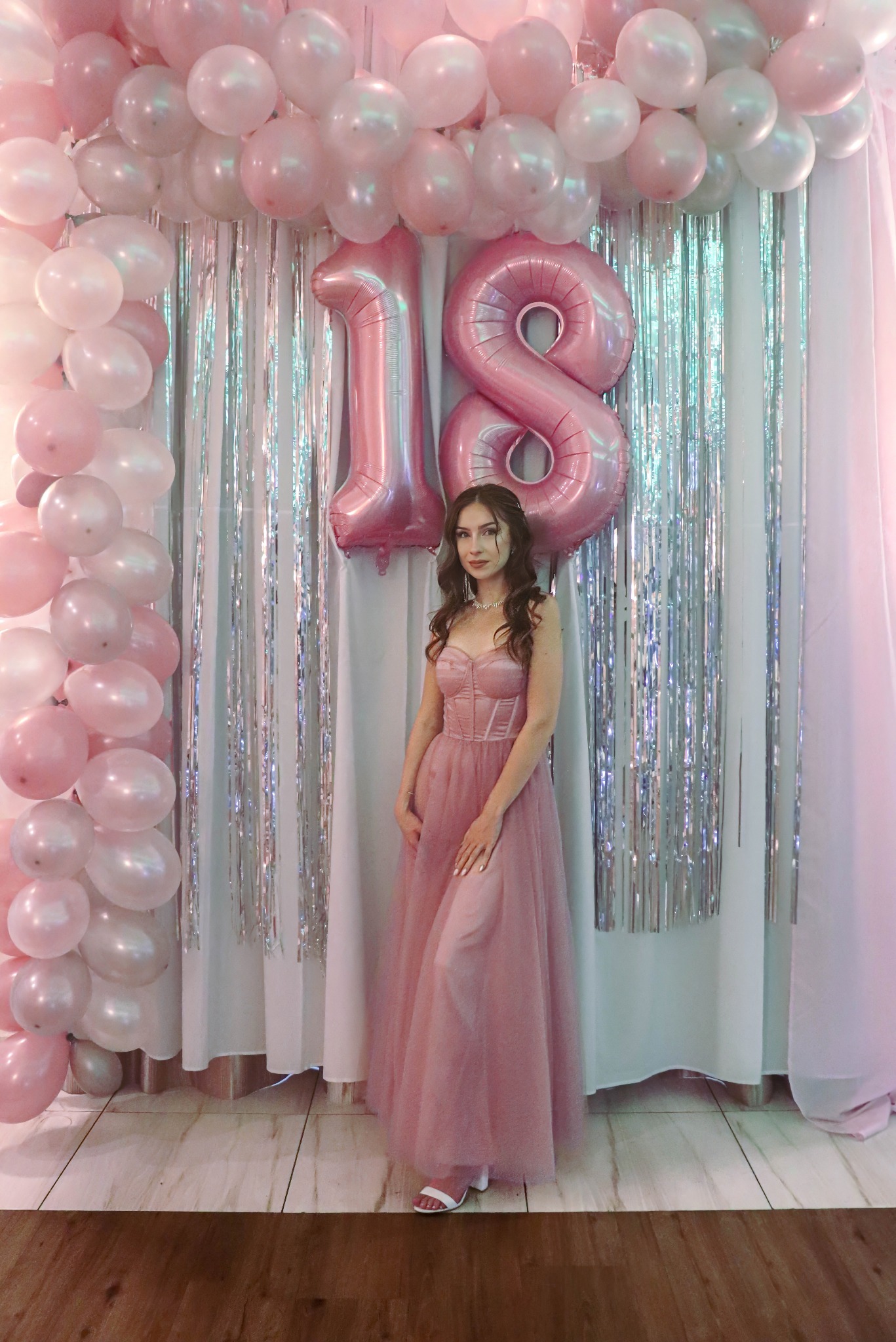 Uśmiechnięta osiemnastolatka w pudrowej sukni, pozuje na tle dekoracji urodzinowej z balonów i srebrnych frędzli. Romantyczna atmosfera, delikatne światło.