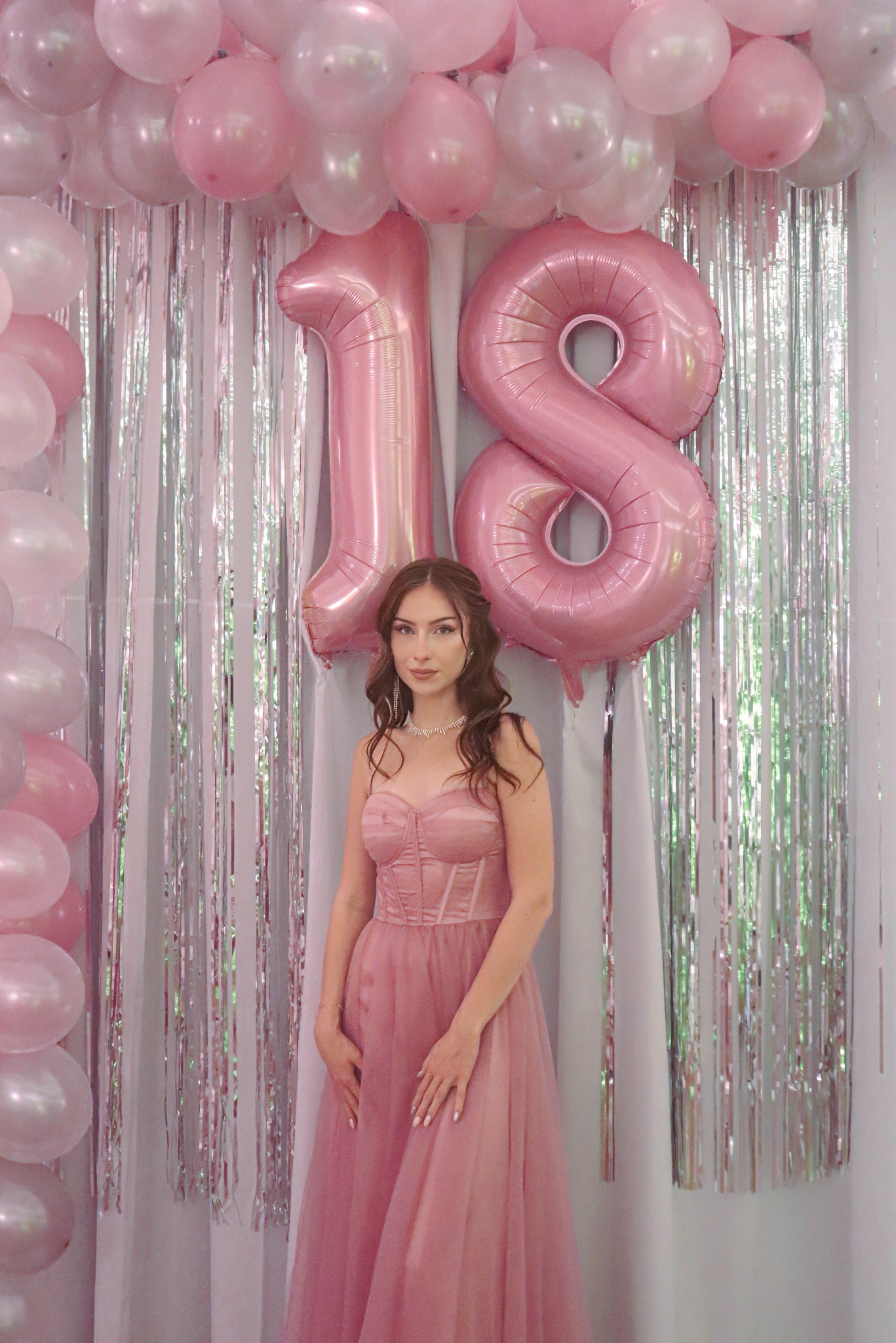 Młoda kobieta w różowej sukni pozuje na tle dekoracji urodzinowych z balonów w kształcie liczby 18 i srebrnych frędzli. Romantyczna sceneria, delikatne oświetlenie.