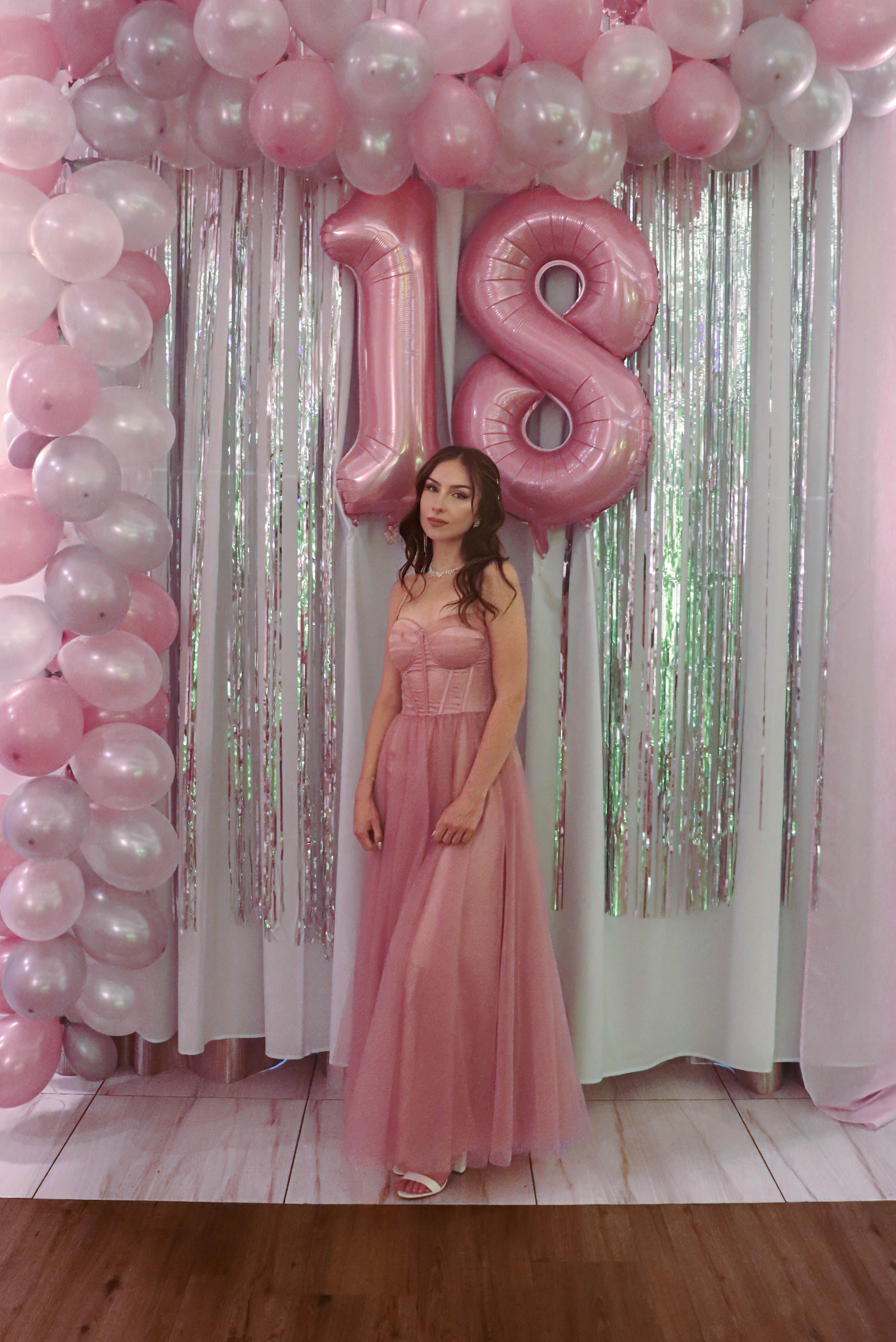 Uśmiechnięta młoda kobieta w różowej sukni pozuje na tle dekoracji urodzinowych z balonami i błyszczącymi frędzlami. Balony układają się w liczbę 18.