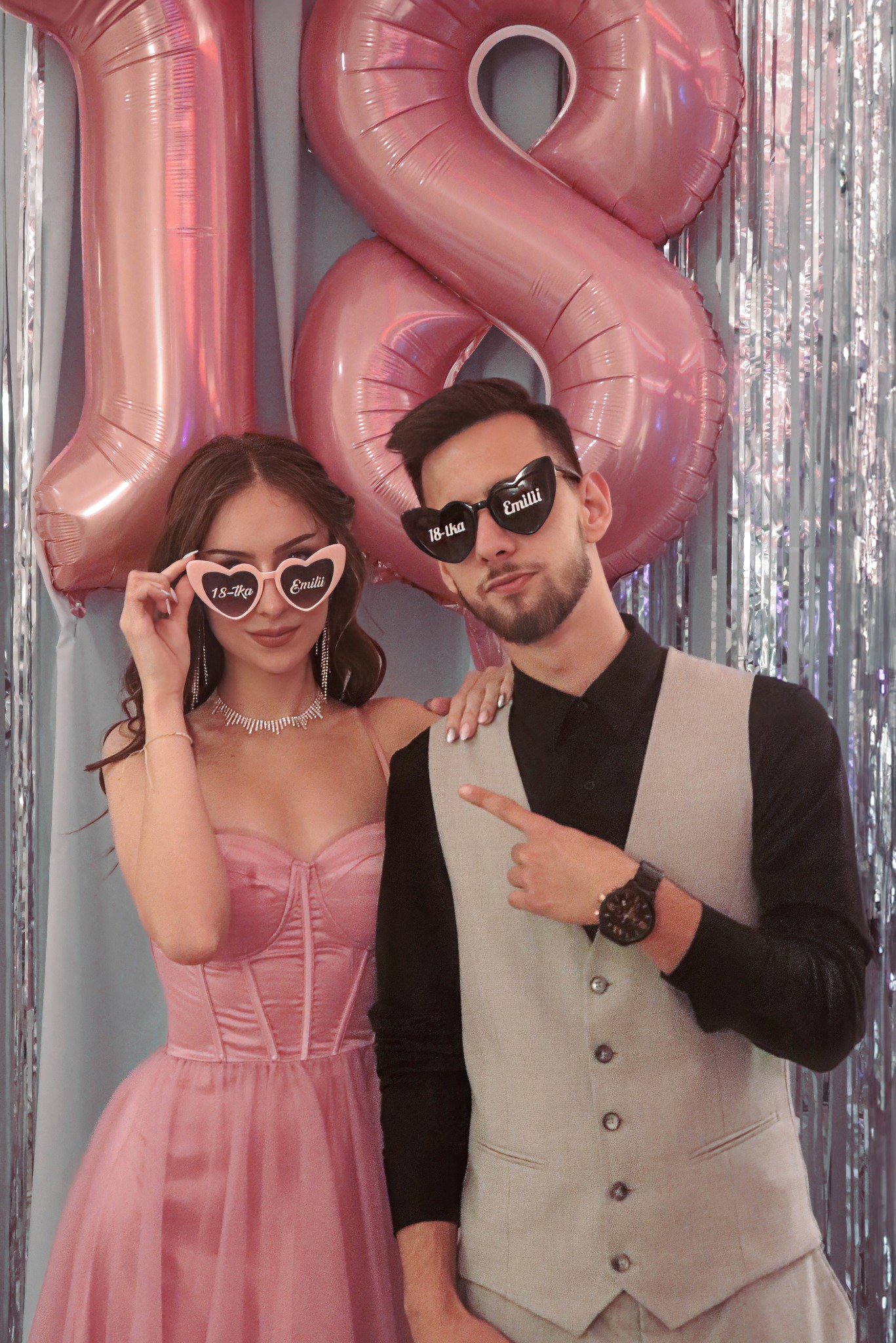 Uśmiechnięta para w okularach w kształcie serc z napisem '18-tka Emilii' pozuje na tle srebrnych frędzli i różowych balonów w kształcie cyfry 18.