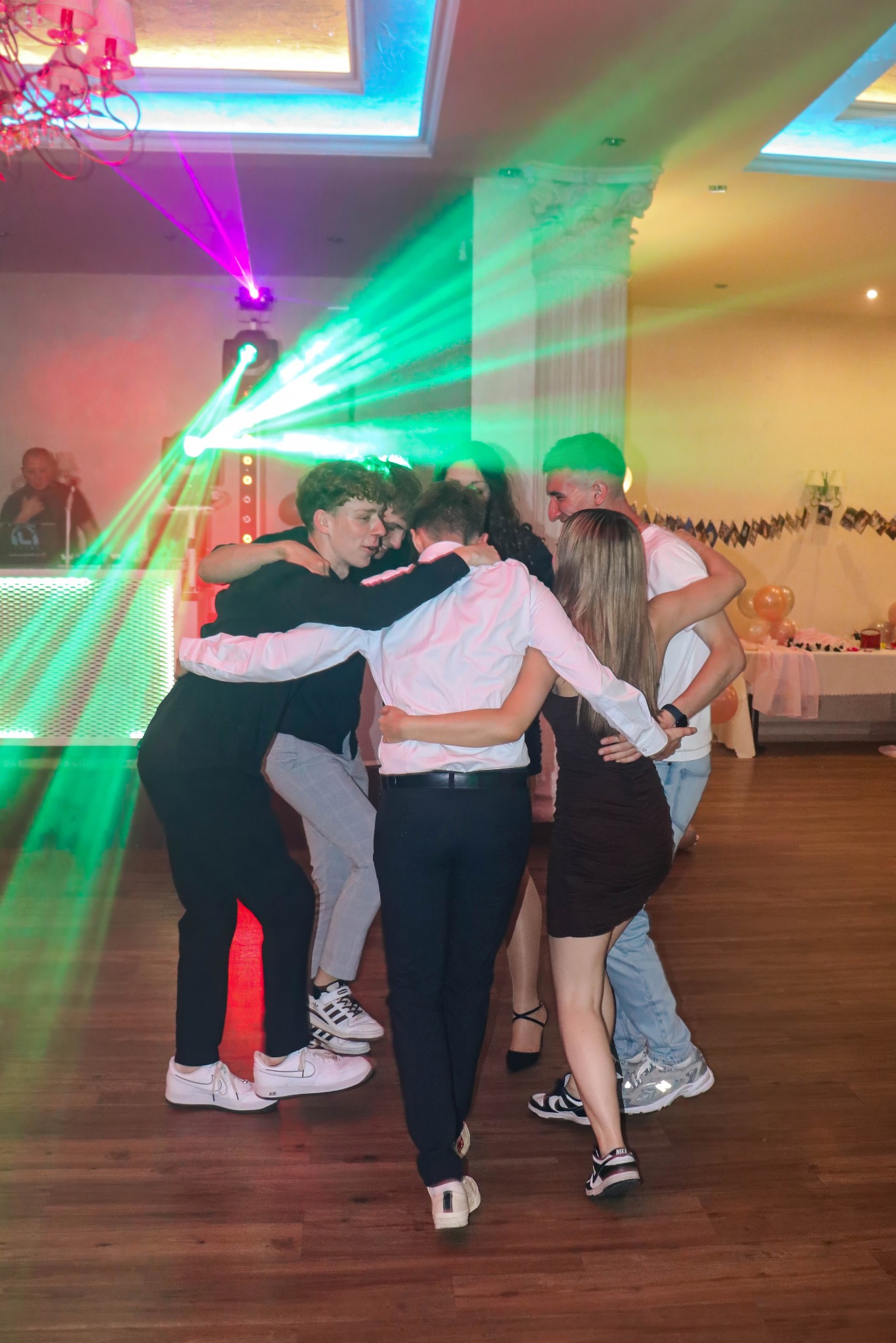 Grupa młodych ludzi w objęciach na parkiecie, oświetlona kolorowymi laserami. W tle DJ na stanowisku i dekoracje imprezowe.