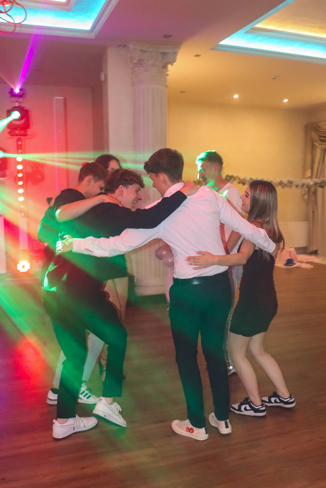 Grupa młodych ludzi tańczy w kręgu na parkiecie, oświetlona kolorowymi światłami dyskotekowymi. Widoczne kolumny i dekoracje sali balowej.
