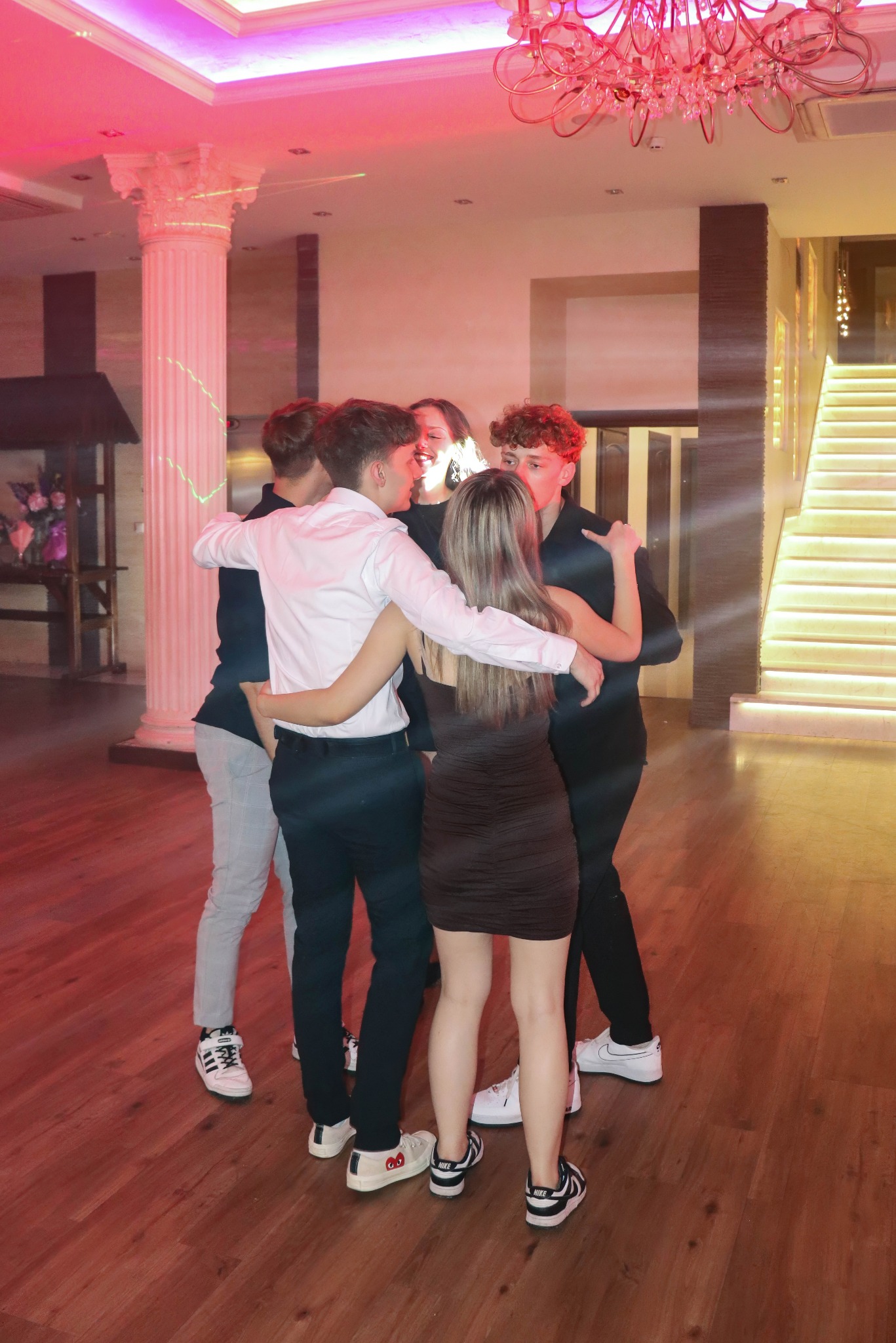 Grupa młodych ludzi w objęciach na parkiecie, kolumna w tle, oświetlenie imprezowe. Uczestnicy wydarzenia w swobodnych strojach, widoczne buty sportowe.