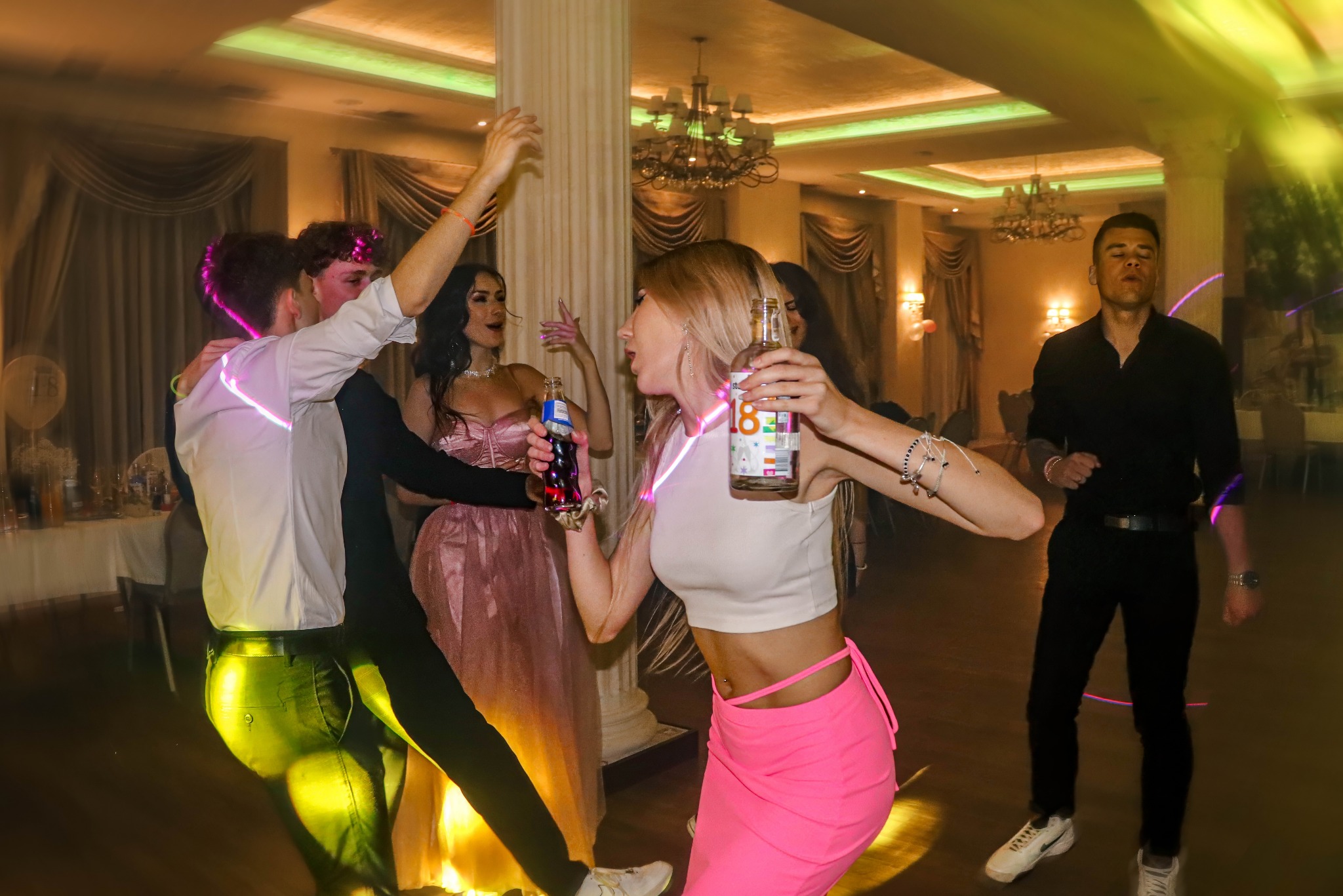 Roztańczona grupa osób na sali balowej podczas imprezy. Kobieta w różowej spódnicy trzyma butelkę z napojem. W tle kolumny i dekoracje.