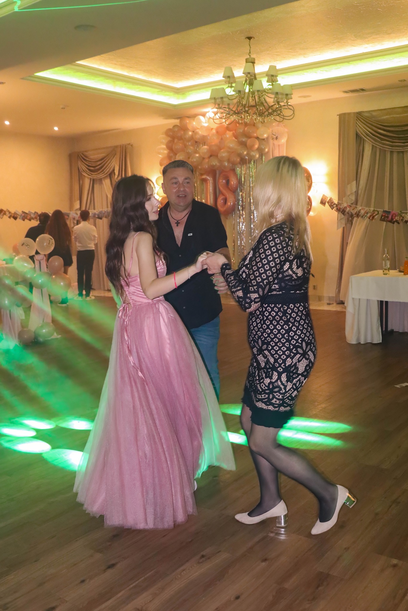 Uczestnicy imprezy tańczą na parkiecie. W tle dekoracje balonowe i oświetlenie sceniczne. Dziewczyna w różowej sukni, mężczyzna w czarnej koszuli i kobieta w czarnej sukience.