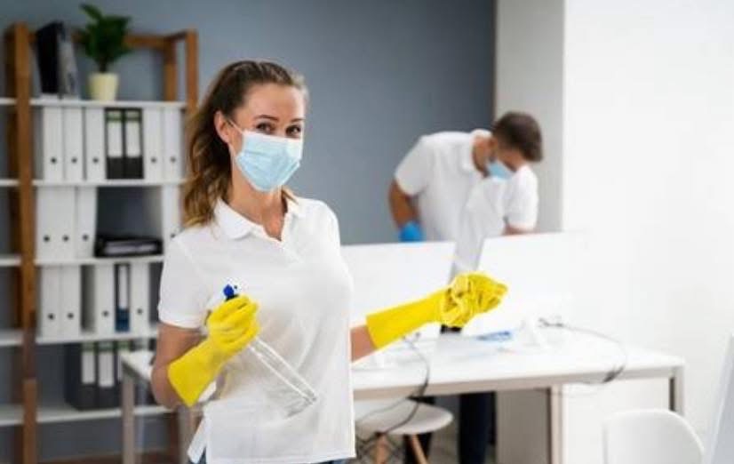 Pracownicy w maseczkach i rękawicach sprzątają biuro. Kobieta z rozpylaczem na pierwszym planie, w tle mężczyzna przy biurku. Półki z segregatorami w tle.