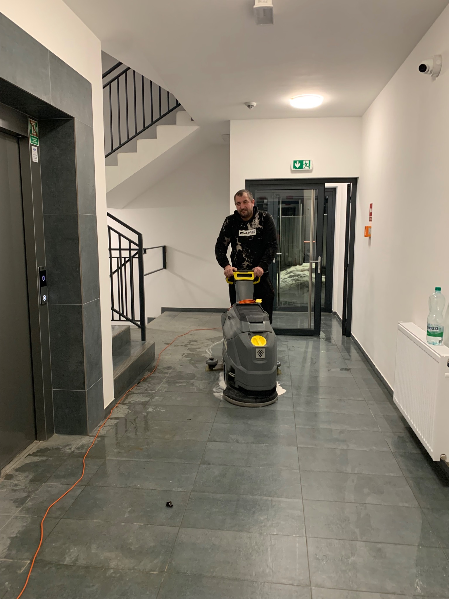 Pracownik w czarnej bluzie czyści posadzkę w korytarzu budynku za pomocą szorowarki. Widoczne płytki podłogowe, winda, schody i butelka wody mineralnej.