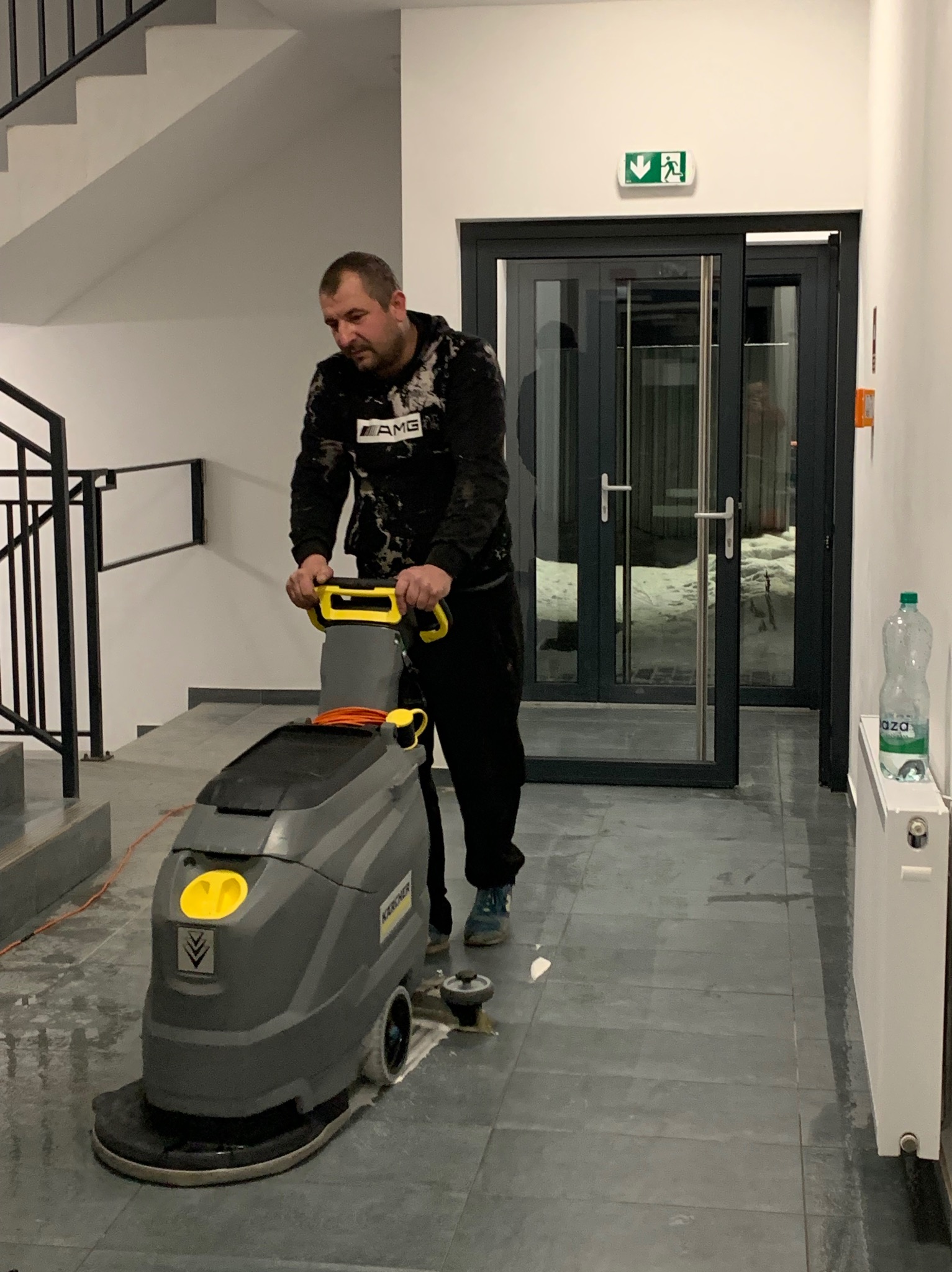 Pracownik w czarnej bluzie AMG czyści podłogę w budynku za pomocą szorowarki Karcher. Widoczny znak ewakuacyjny i butelka wody. Szare płytki podłogowe.