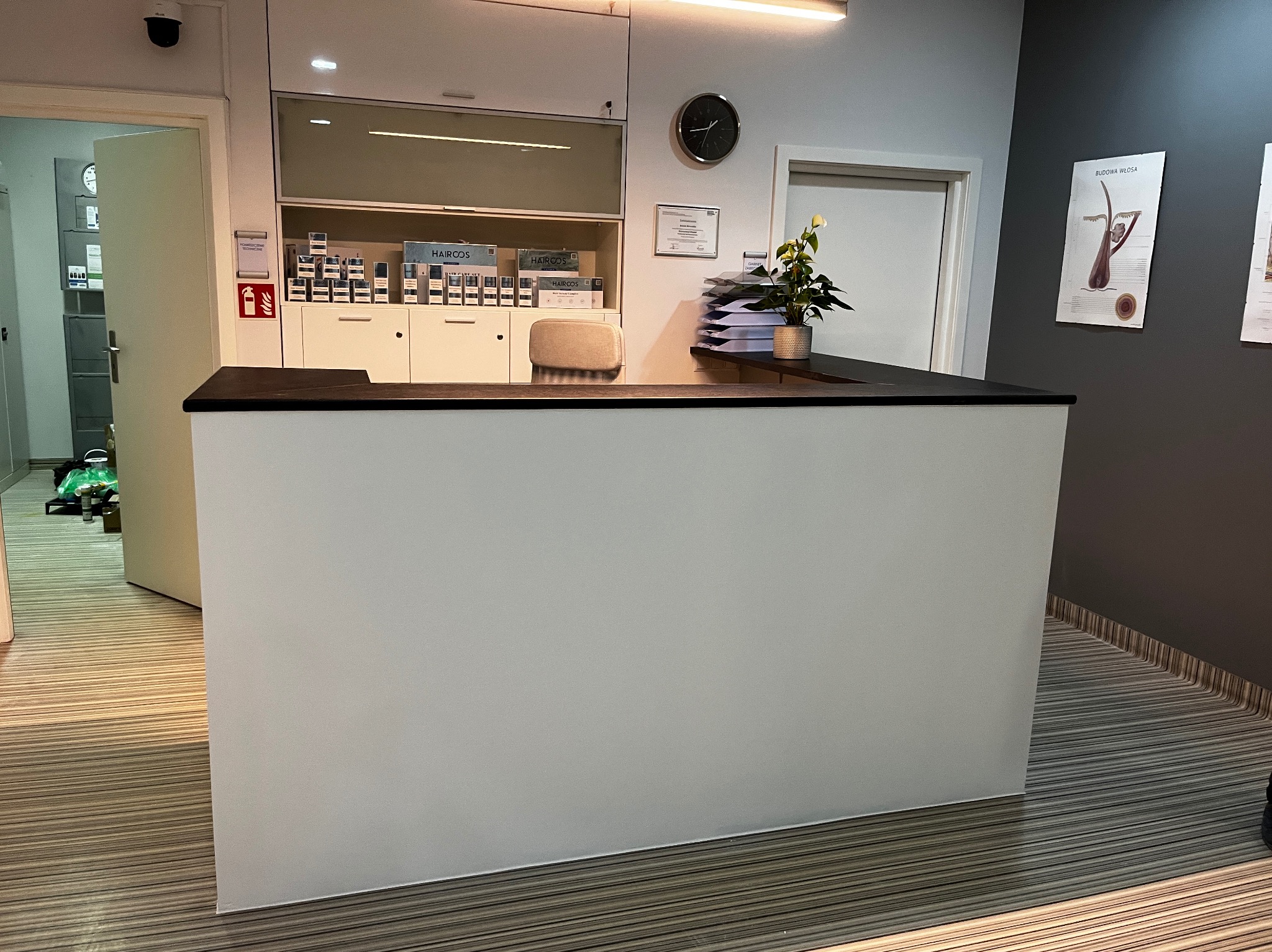 Nowoczesna recepcja biurowa z białym frontem i czarnym blatem, widok na zaplecze z szafkami i produktami. Minimalistyczny design, neutralna kolorystyka, estetyczne wykończenie.