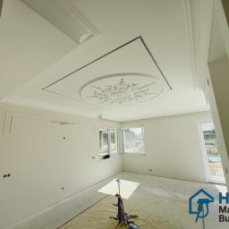 HLK MASTER BUD OLEKSII HLADKOV - Wykończone wnętrze z dekoracyjnym sufitem podwieszanym z gipsowymi zdobieniami. Widoczne przygotowania do dalszych prac wykończeniowych, zabezpieczona podłoga.