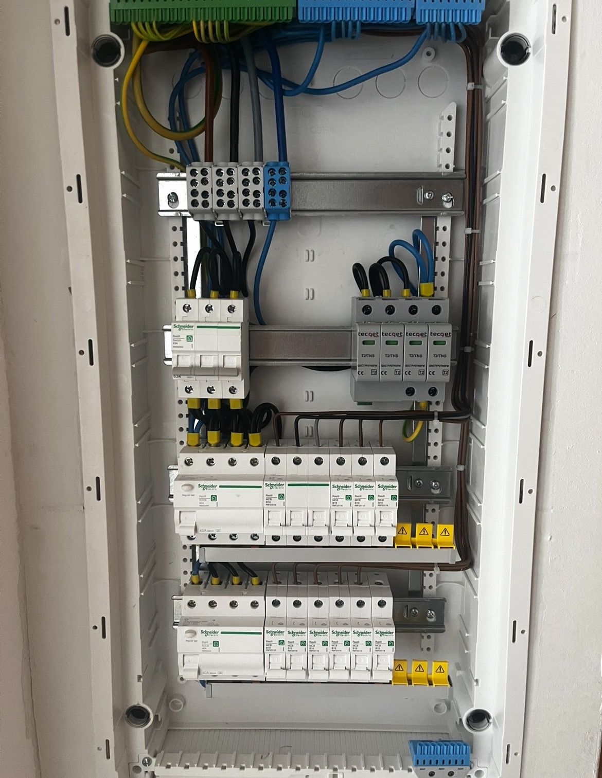 Rozdzielnica elektryczna w białej obudowie z widocznymi bezpiecznikami, przewodami i elementami montażowymi. Widok z bliska na zainstalowane komponenty.