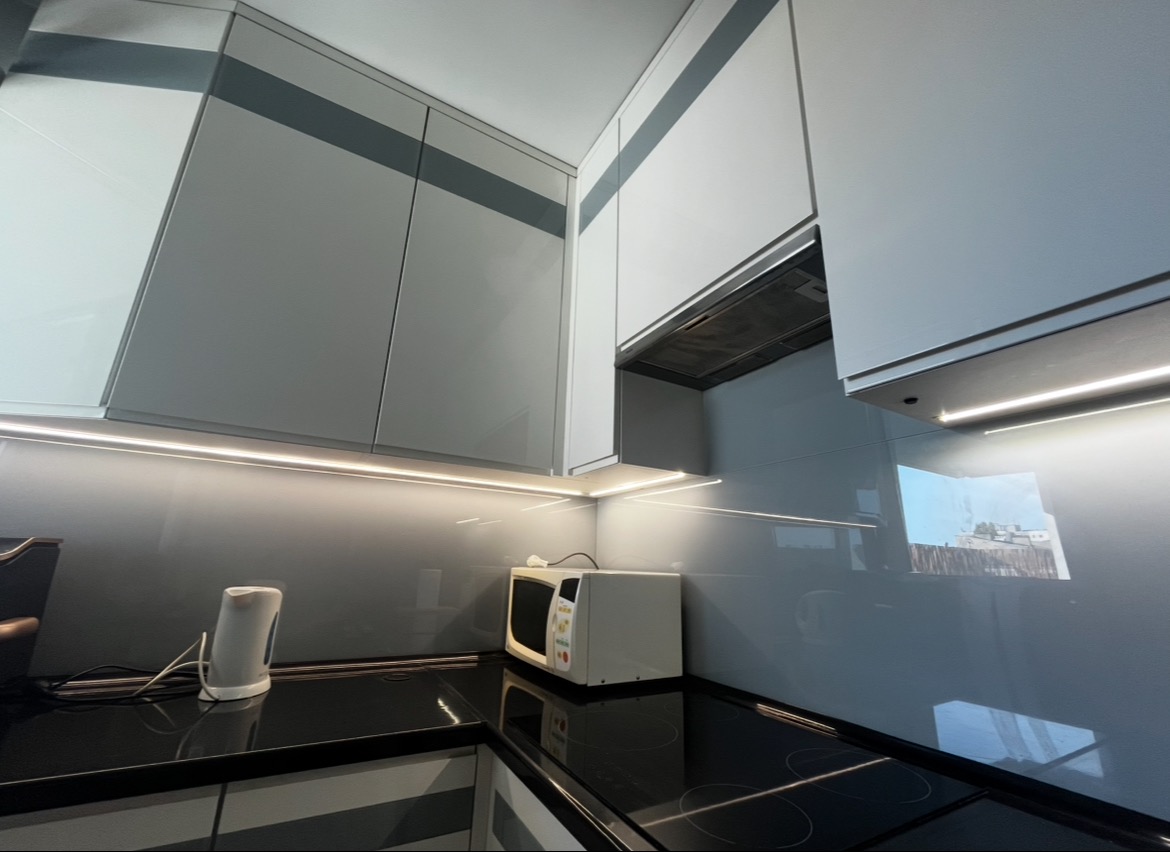 Nowoczesna kuchnia z szarymi szafkami i oświetleniem LED pod szafkami, odbijającym się w błyszczącej powierzchni blatu. Widoczny okap i sprzęt AGD.