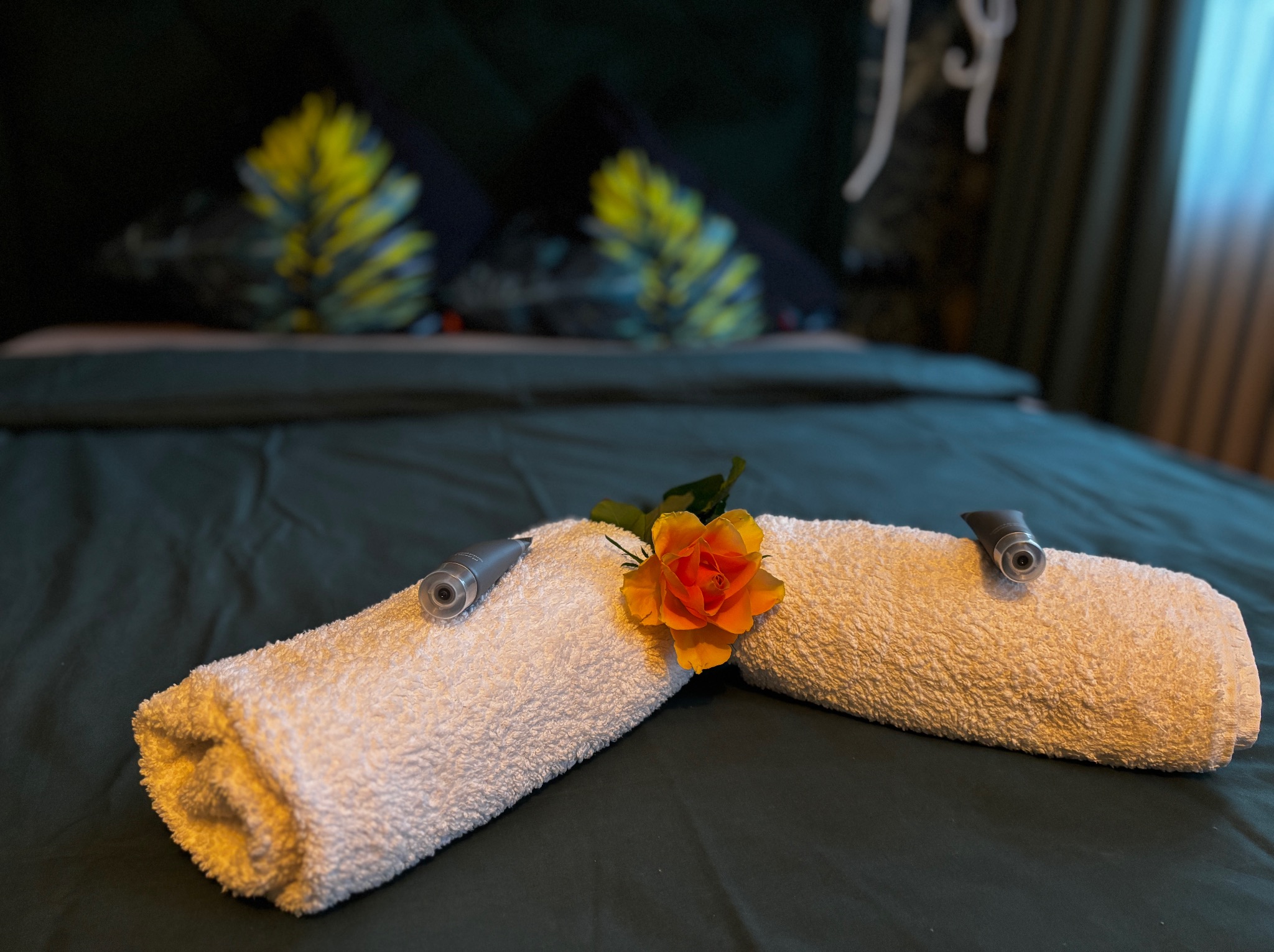 Ułożony ręcznik na łóżku z kwiatem i kosmetykami, ciemnozielona pościel, dekoracyjne poduszki w tle. Przygotowanie pokoju hotelowego.