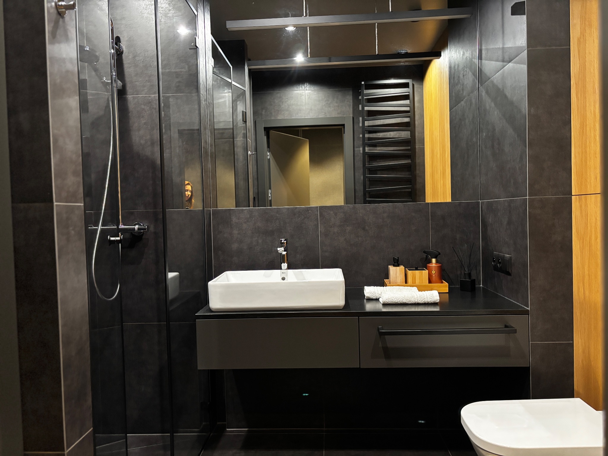 Nowoczesna łazienka w ciemnej tonacji z kabiną prysznicową, umywalką nablatową i dużym lustrem. Widoczne detale wykończenia i oświetlenie.