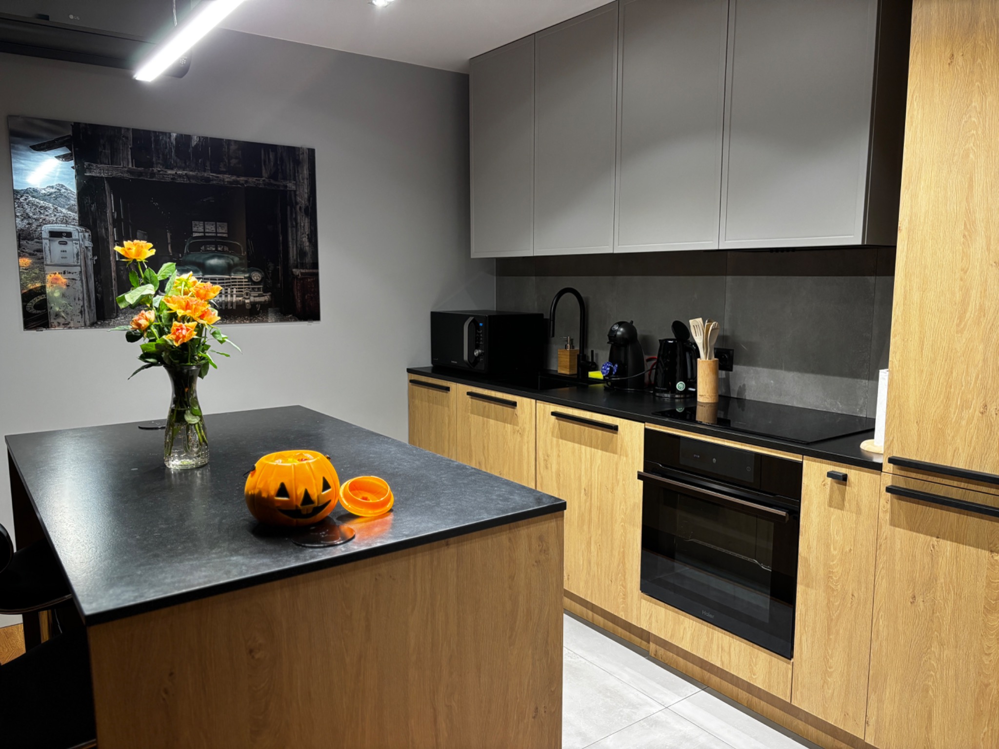 Nowoczesna kuchnia z wyspą, dekoracją Halloween (dynia) i bukietem kwiatów. Jasne drewniane szafki, szare ściany i czarne blaty. Minimalistyczny design.