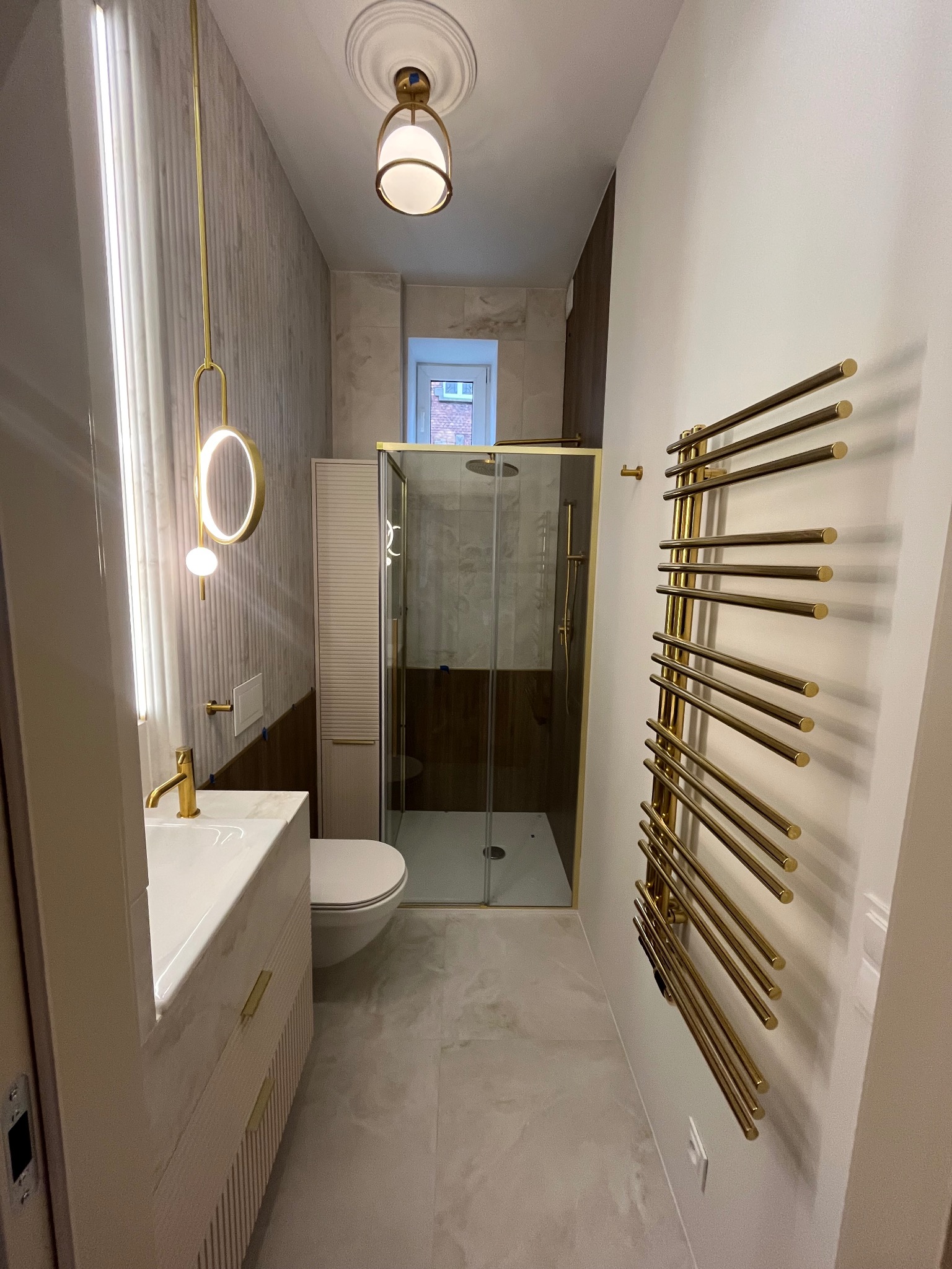 Nowoczesna łazienka po remoncie: kabina prysznicowa ze złotymi ramami, umywalka z marmurowym blatem, złoty grzejnik drabinkowy i designerskie oświetlenie.