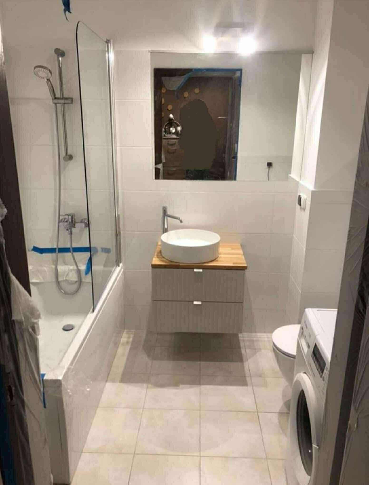 Jasna łazienka po remoncie: białe płytki, umywalka nablatowa na drewnianym blacie, kabina prysznicowa. Widoczna pralka i lustro. Minimalistyczny design.