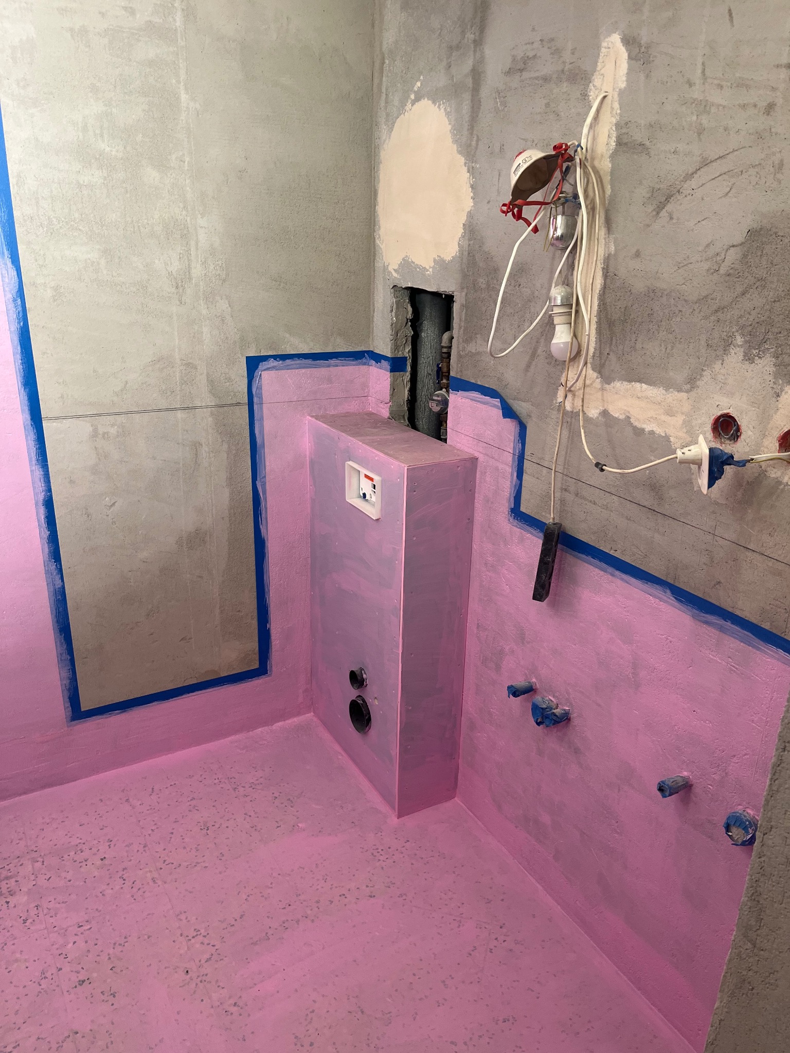 Hydroizolacja w łazience w trakcie remontu: różowa powłoka na podłodze i ścianach, zabezpieczone rury, surowy beton, niebieska taśma malarska.