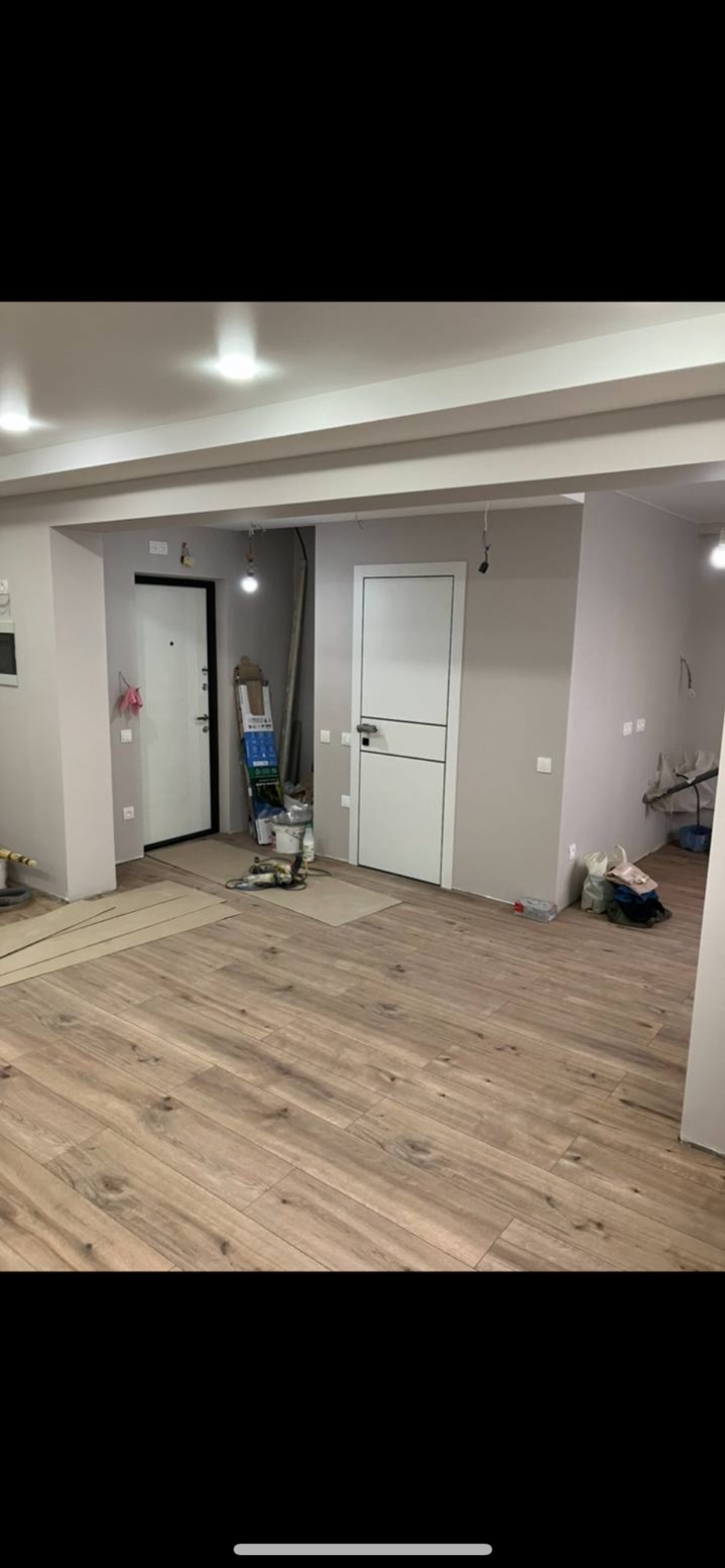 Jasne panele podłogowe w nowym mieszkaniu, świeżo po remoncie. Widoczne dwie białe, nowoczesne drzwi i ściany pomalowane na szaro. W tle drabina i narzędzia.