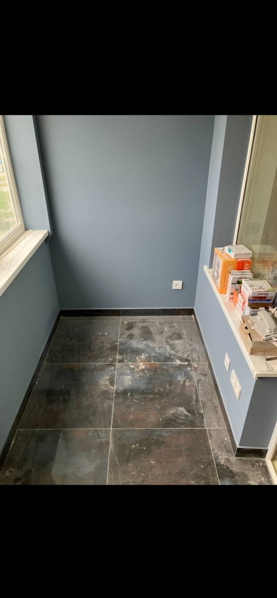 Balkon po malowaniu ścian na szaro, z ciemną, zabrudzoną podłogą z płytek. Widoczne gniazdka elektryczne i materiały budowlane na parapecie.