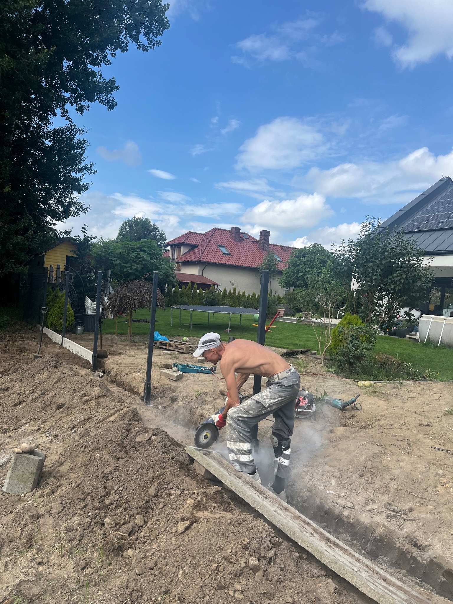 Pracownik tnie betonową płytę szlifierką kątową podczas montażu ogrodzenia. Widoczne słupki ogrodzeniowe, wykop i domy w tle. Praca w toku.